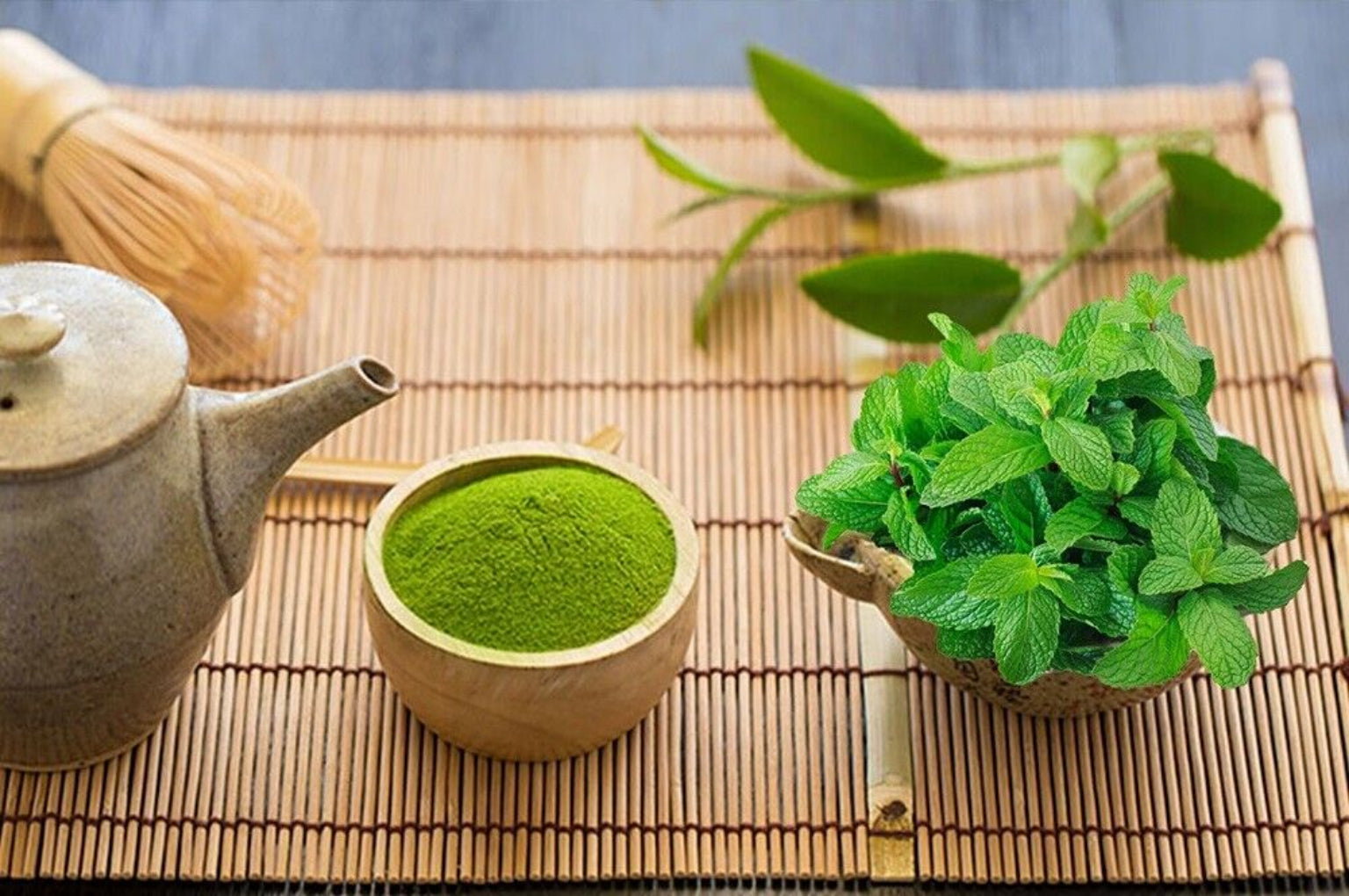 A wooden bowl filled with green powder next to fresh peppermint leaves on a bamboo mat, with a ceramic tea set in the background.